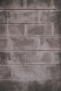 A concrete block wall with a rough texture, featuring rectangular blocks stacked in a staggered pattern. The surface appears weathered, with some areas showing darker stains or marks.