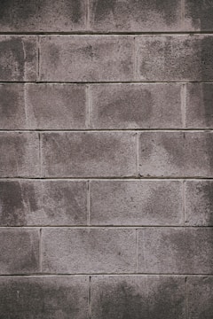 A concrete block wall with a rough texture, featuring rectangular blocks stacked in a staggered pattern. The surface appears weathered, with some areas showing darker stains or marks.