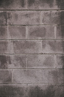 A concrete block wall with a rough texture, featuring rectangular blocks stacked in a staggered pattern. The surface appears weathered, with some areas showing darker stains or marks.