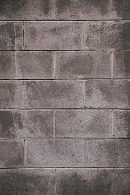 A concrete block wall with a rough texture, featuring rectangular blocks stacked in a staggered pattern. The surface appears weathered, with some areas showing darker stains or marks.