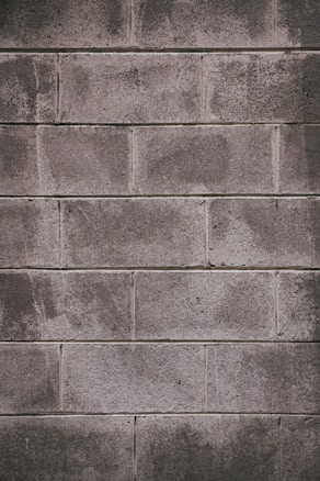 A concrete block wall with a rough texture, featuring rectangular blocks stacked in a staggered pattern. The surface appears weathered, with some areas showing darker stains or marks.