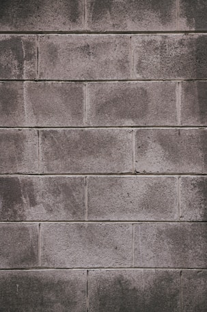 A concrete block wall with a rough texture, featuring rectangular blocks stacked in a staggered pattern. The surface appears weathered, with some areas showing darker stains or marks.
