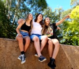 Three young people are sitting on a stone wall, smiling and engaging in a moment of laughter and camaraderie. They have their arms around each other, with the person on the right gesturing or pointing towards something in the distance. The background is filled with lush green trees, suggesting a sunny day spent outdoors.