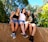 Three young people are sitting on a stone wall, smiling and engaging in a moment of laughter and camaraderie. They have their arms around each other, with the person on the right gesturing or pointing towards something in the distance. The background is filled with lush green trees, suggesting a sunny day spent outdoors.