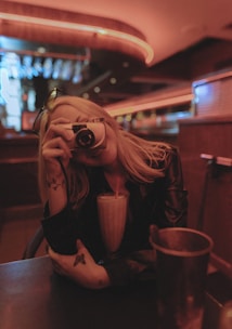 A person with long hair holds a camera up to their face as if taking a picture. They are sitting at a dimly-lit booth with a milkshake in front of them, featuring a red straw. The setting has a relaxed, vintage feel with warm lighting and visible ceiling light fixtures.