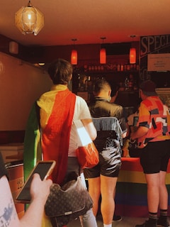 A group of people stand at a bar counter decorated with a rainbow flag. One person is draped in a rainbow-colored garment, while others wear colorful, casual clothing. Above, several red pendant lights illuminate the scene, adding warmth to the atmosphere.