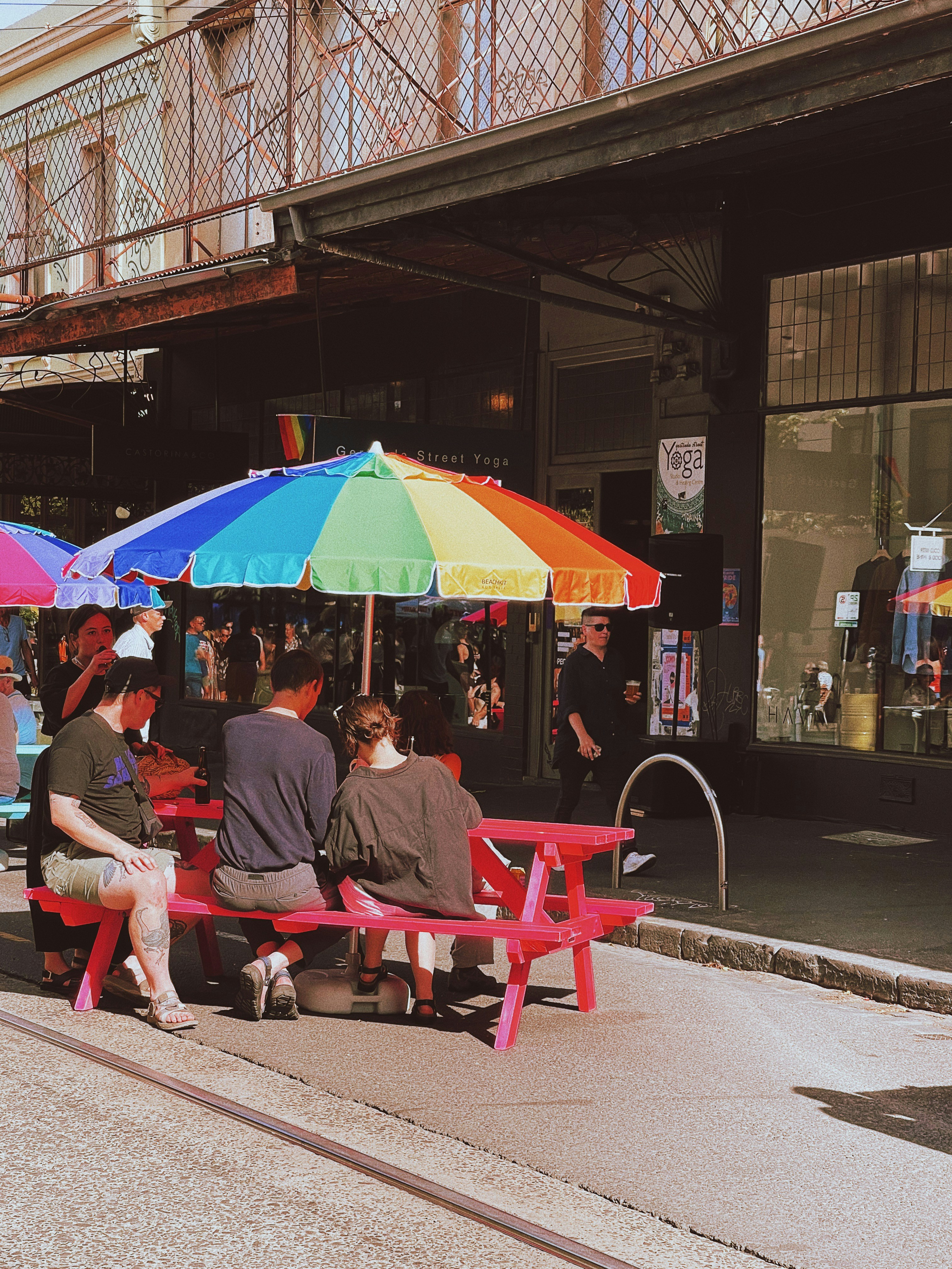 Three people at a pink picnic table under a rainbow umbrella at a sidewalk cafe.