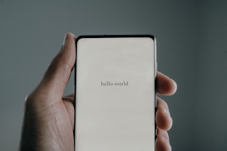 A hand holds a smartphone displaying the text 'hello world' on the screen. The background is a muted gray, and the lighting creates a soft shadow around the hand.