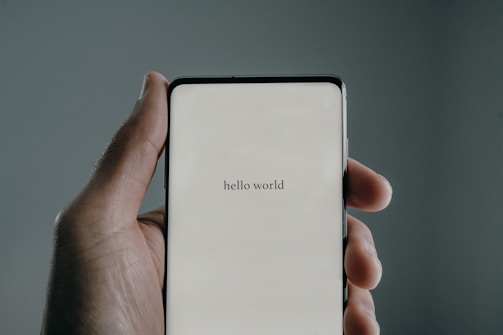 A hand holds a smartphone displaying the text 'hello world' on the screen. The background is a muted gray, and the lighting creates a soft shadow around the hand.