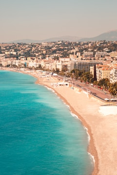 A picturesque coastal cityscape with a vibrant blue sea meeting a sandy shoreline. The city is densely packed with multi-story buildings that stretch along the coast, backed by lush green hills. The beach appears calm, and the atmosphere is sunny and inviting.