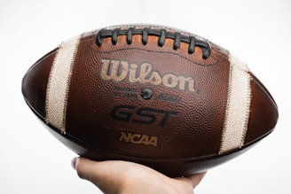 A close-up of a Wilson GST NCAA football held in a person's hand. The football has a textured brown surface with white stripes and black laces. The brand name Wilson is prominently displayed along with the GST and NCAA logos.