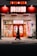 A person walks past a brightly lit karaoke establishment with prominent red signage and illuminated posters in Japanese. The scene takes place at night, with the warm glow of lights enhancing the contrast against the dark street.