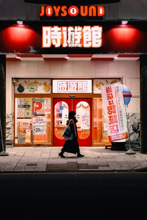 A person walks past a brightly lit karaoke establishment with prominent red signage and illuminated posters in Japanese. The scene takes place at night, with the warm glow of lights enhancing the contrast against the dark street.
