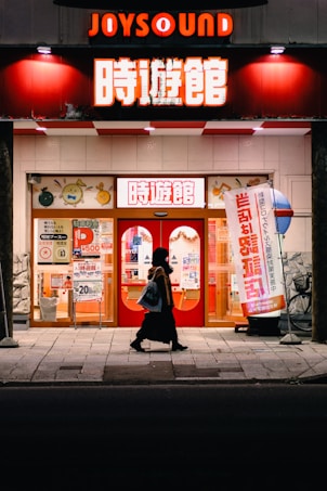 A person walks past a brightly lit karaoke establishment with prominent red signage and illuminated posters in Japanese. The scene takes place at night, with the warm glow of lights enhancing the contrast against the dark street.