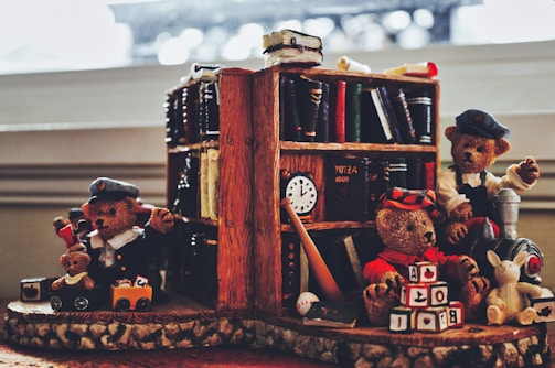 Three teddy bears are positioned around a wooden bookshelf filled with books. One teddy bear in a conductor uniform holds a toy train on tracks. Another teddy bear is dressed casually and sits on a toy train with a stuffed rabbit beside it. The third teddy bear wears a red outfit and is playing with alphabet blocks. A toy baseball and book are in the foreground.