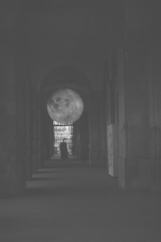 A dimly lit corridor features a large spherical object resembling the moon, suspended in the center. The corridor is framed by tall archways on either side, leading to a brighter area in the distance. The atmospheric lighting adds mystery to the scene.