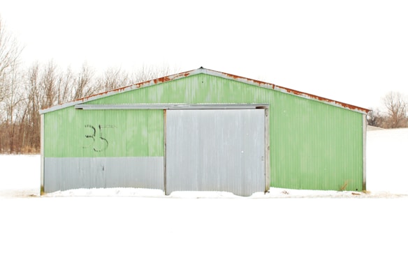 A metal building with a corrugated surface painted in green and gray stands against a snow-covered landscape. The number '35' is marked in black on the green upper part of the structure. Bare trees are visible in the background, providing a stark contrast to the white snow blanketing the ground.