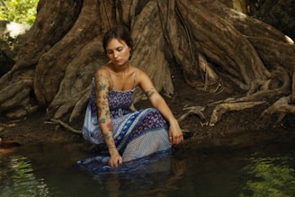 A woman with tattoos sits beside a tranquil body of water, surrounded by large, twisted tree roots. She wears a patterned blue and white dress and appears thoughtful, with her hand gently touching the water's surface.