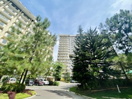 Tall residential buildings surrounded by lush green trees under a bright blue sky. Several parked cars are visible near the buildings, and the area appears well-maintained with green lawns and flower beds.