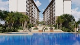 A large, attractive residential complex with twin, multi-story buildings frames a central pool area. Lush greenery, including tall palm-like trees and shrubs, surrounds the pool, creating a resort-like atmosphere. People are casually strolling around the pool area, and there are outdoor seating structures in the distance.