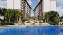 A large, attractive residential complex with twin, multi-story buildings frames a central pool area. Lush greenery, including tall palm-like trees and shrubs, surrounds the pool, creating a resort-like atmosphere. People are casually strolling around the pool area, and there are outdoor seating structures in the distance.