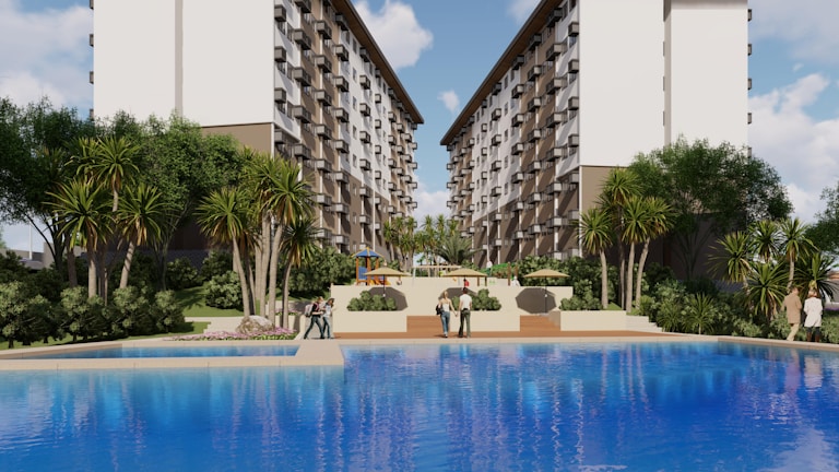 A large, attractive residential complex with twin, multi-story buildings frames a central pool area. Lush greenery, including tall palm-like trees and shrubs, surrounds the pool, creating a resort-like atmosphere. People are casually strolling around the pool area, and there are outdoor seating structures in the distance.