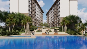 A large, attractive residential complex with twin, multi-story buildings frames a central pool area. Lush greenery, including tall palm-like trees and shrubs, surrounds the pool, creating a resort-like atmosphere. People are casually strolling around the pool area, and there are outdoor seating structures in the distance.