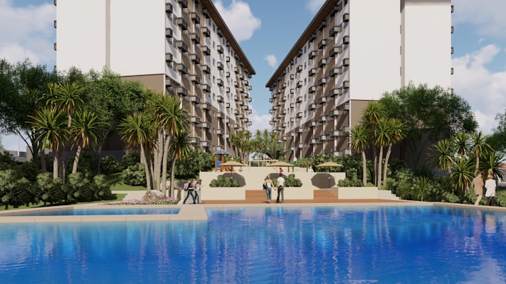 A large, attractive residential complex with twin, multi-story buildings frames a central pool area. Lush greenery, including tall palm-like trees and shrubs, surrounds the pool, creating a resort-like atmosphere. People are casually strolling around the pool area, and there are outdoor seating structures in the distance.