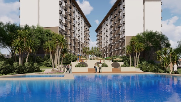 A large, attractive residential complex with twin, multi-story buildings frames a central pool area. Lush greenery, including tall palm-like trees and shrubs, surrounds the pool, creating a resort-like atmosphere. People are casually strolling around the pool area, and there are outdoor seating structures in the distance.