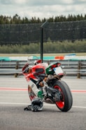 A high-performance motorcycle with an Italian flag-themed color scheme is parked on a racetrack. It has a sleek, aerodynamic design with visible branding. A black helmet is placed on the ground next to the motorcycle. The background includes a section of the racetrack and a fence, with trees visible in the distance under a cloudy sky.