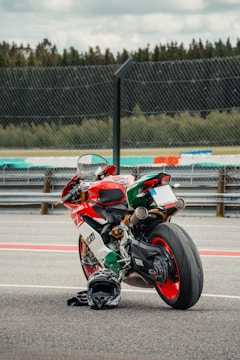 A high-performance motorcycle with an Italian flag-themed color scheme is parked on a racetrack. It has a sleek, aerodynamic design with visible branding. A black helmet is placed on the ground next to the motorcycle. The background includes a section of the racetrack and a fence, with trees visible in the distance under a cloudy sky.
