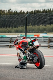 A high-performance motorcycle with an Italian flag-themed color scheme is parked on a racetrack. It has a sleek, aerodynamic design with visible branding. A black helmet is placed on the ground next to the motorcycle. The background includes a section of the racetrack and a fence, with trees visible in the distance under a cloudy sky.