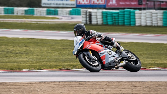 A motorcycle racer leans into a turn on a racetrack, showcasing dynamic speed and control. The rider wears a full racing suit and helmet, and the bike is branded with vibrant graphics.