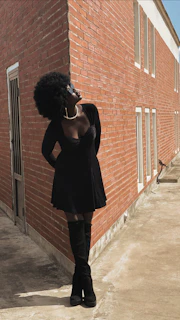 A person with an afro hairstyle is standing against a brick wall, dressed in a black outfit with sunglasses and a pearl necklace. The wall has multiple windows and a door, casting shadows on the ground.