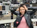 A smiling woman holding a sleek Bluetooth speaker with neon-lit shelves behind her.