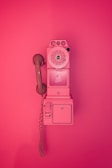 A vintage rotary payphone mounted on a wall, entirely in shades of pink. The phone has a round dial with a handset attached by a curly cord and features a nostalgic design reminiscent of mid-20th century telecommunication devices.