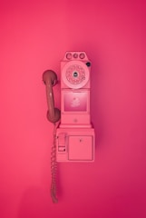 A vintage rotary payphone mounted on a wall, entirely in shades of pink. The phone has a round dial with a handset attached by a curly cord and features a nostalgic design reminiscent of mid-20th century telecommunication devices.