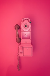 A vintage rotary payphone mounted on a wall, entirely in shades of pink. The phone has a round dial with a handset attached by a curly cord and features a nostalgic design reminiscent of mid-20th century telecommunication devices.