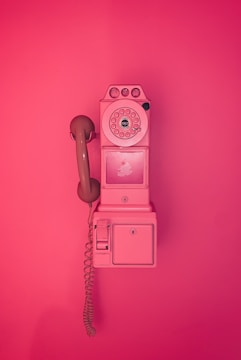 A vintage rotary payphone mounted on a wall, entirely in shades of pink. The phone has a round dial with a handset attached by a curly cord and features a nostalgic design reminiscent of mid-20th century telecommunication devices.