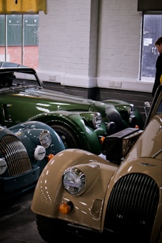 A collection of vintage sports cars with classic, rounded designs. The vehicles are parked indoors, beneath a window with a yellow curtain. The cars feature various colors including green, blue, and beige, with distinctive chrome headlights and grilles. A person stands in the background near the window.