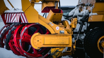 A close-up view of a yellow snow plow machine with red rotating blades. The surface shows signs of wear and there is snow clinging to various parts. The background is bright and overcast, indicating a snowy environment.