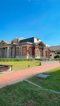 A historic building with red brick and ornate architectural details stands amidst a well-maintained grassy area. The building features a dark roof and tall columns at the entrance, with a clear blue sky overhead. A paved path and benches are visible in the foreground.