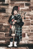 A person dressed in traditional Scottish attire is playing bagpipes while standing against a stone wall. They are wearing a patterned kilt, a dark jacket with silver buttons, and a fur hat. The setting appears to be historical, conveying a sense of heritage and tradition.