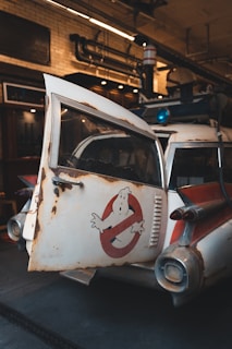 A vintage car with a weathered and rusty exterior is parked indoors. The vehicle features a distinctive logo of a white figure on a red circle and slash on the door. The interior environment has industrial elements like exposed pipes and brick walls.