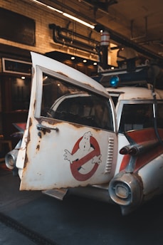A vintage car with a weathered and rusty exterior is parked indoors. The vehicle features a distinctive logo of a white figure on a red circle and slash on the door. The interior environment has industrial elements like exposed pipes and brick walls.