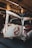 A vintage car with a weathered and rusty exterior is parked indoors. The vehicle features a distinctive logo of a white figure on a red circle and slash on the door. The interior environment has industrial elements like exposed pipes and brick walls.