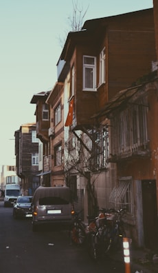 A narrow, residential street lined with older, multi-story buildings featuring wooden facades and small, white-framed windows. A Turkish flag is draped from one of the buildings. Several cars are parked along the street, and bicycles are leaning against the wall. The area appears quiet and somewhat rustic, with a few bare trees visible.