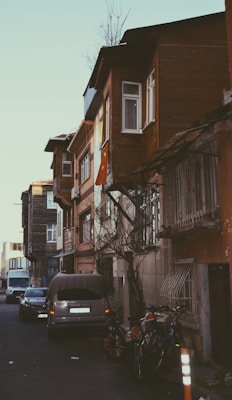 A narrow, residential street lined with older, multi-story buildings featuring wooden facades and small, white-framed windows. A Turkish flag is draped from one of the buildings. Several cars are parked along the street, and bicycles are leaning against the wall. The area appears quiet and somewhat rustic, with a few bare trees visible.