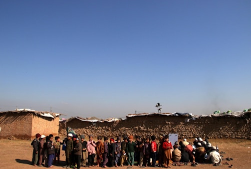A group of children is gathered in an open area with an expansive blue sky above them. The children are dressed in various layers of clothing, suggesting cooler weather. Behind them, there are low, makeshift mud-brick structures, indicating a rural or underdeveloped setting. Some children are standing, while others are seated on the ground, possibly engaged in an activity.