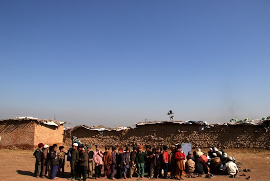 A group of children is gathered in an open area with an expansive blue sky above them. The children are dressed in various layers of clothing, suggesting cooler weather. Behind them, there are low, makeshift mud-brick structures, indicating a rural or underdeveloped setting. Some children are standing, while others are seated on the ground, possibly engaged in an activity.